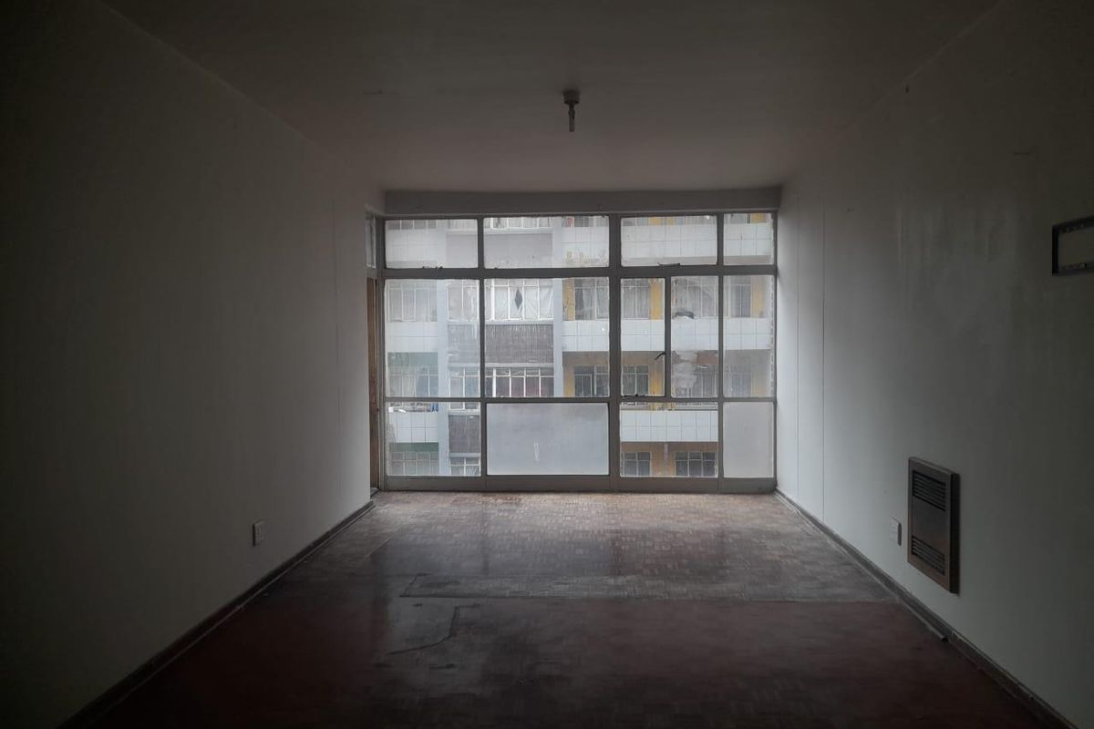 An empty, sunlit living room featuring original parquet flooring, large windows with neighborhood views, and a wall-mounted heater.