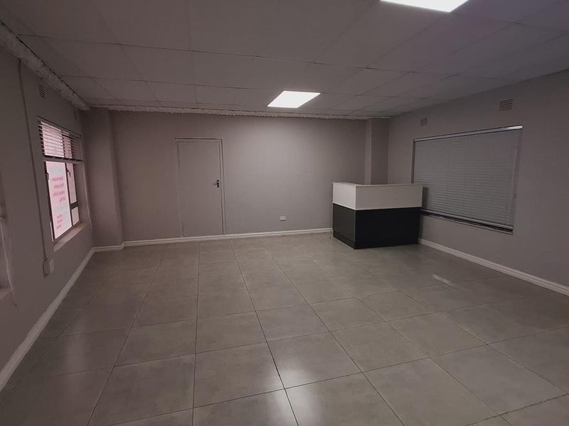 A spacious, empty office area featuring neutral tiled flooring, a built-in reception counter, and recessed ceiling lighting, ready for professional use.