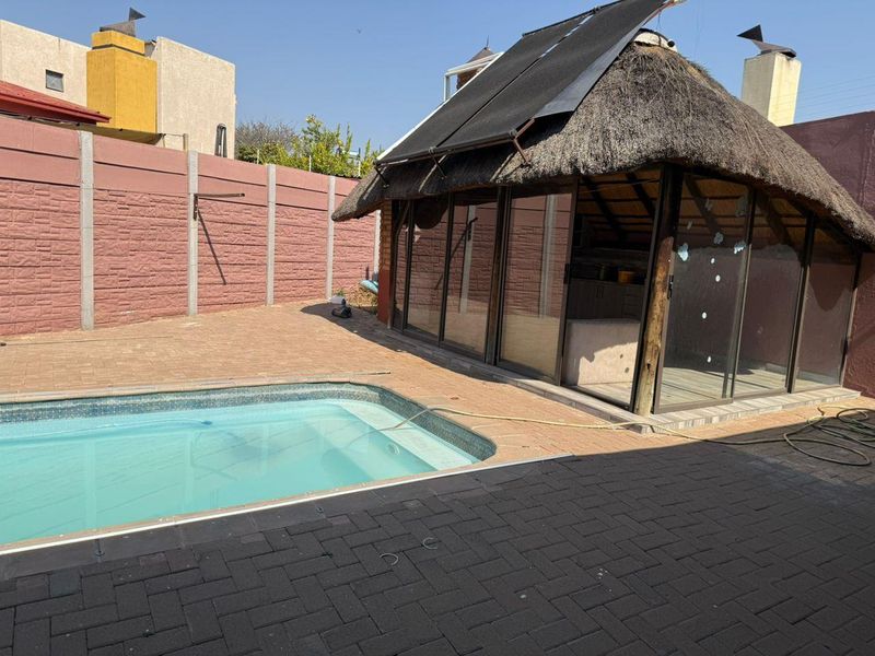Outdoor entertainment area featuring a swimming pool, paved patio, and a thatched lapa structure with sliding glass doors and solar heating panels.