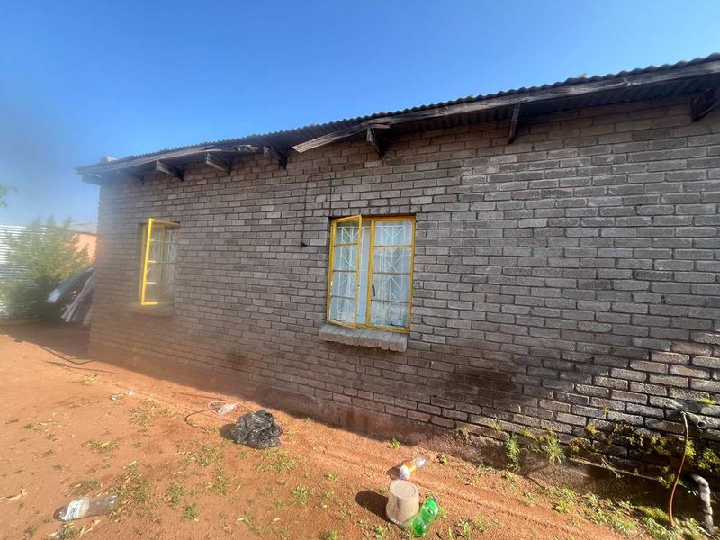 Exterior view of a brick house featuring corrugated iron roofing and steel window frames with security bars, showing signs of significant weathering and maintenance needs.