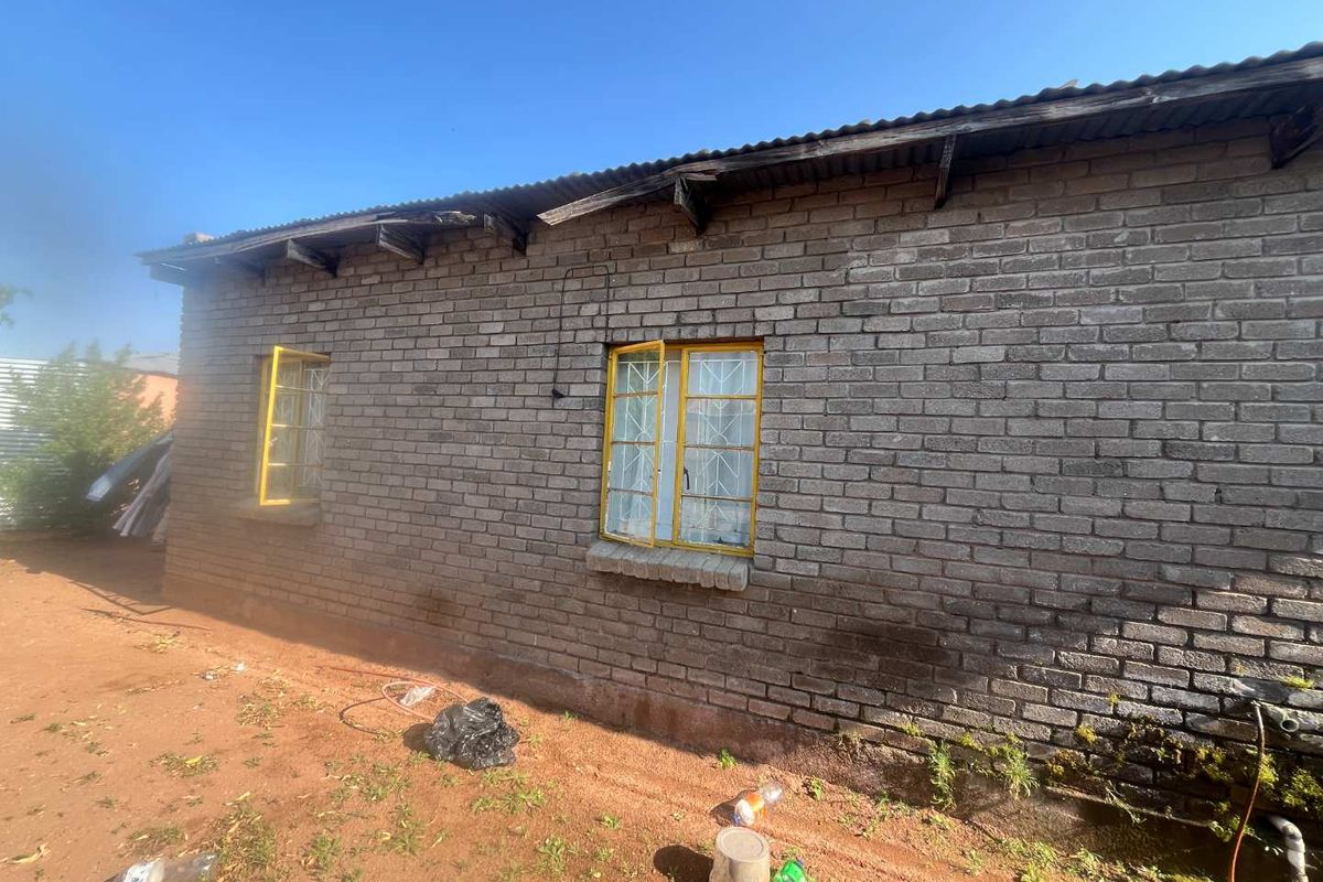 Exterior view of a brick house featuring corrugated iron roofing and steel window frames with security bars, showing signs of significant weathering and maintenance needs.