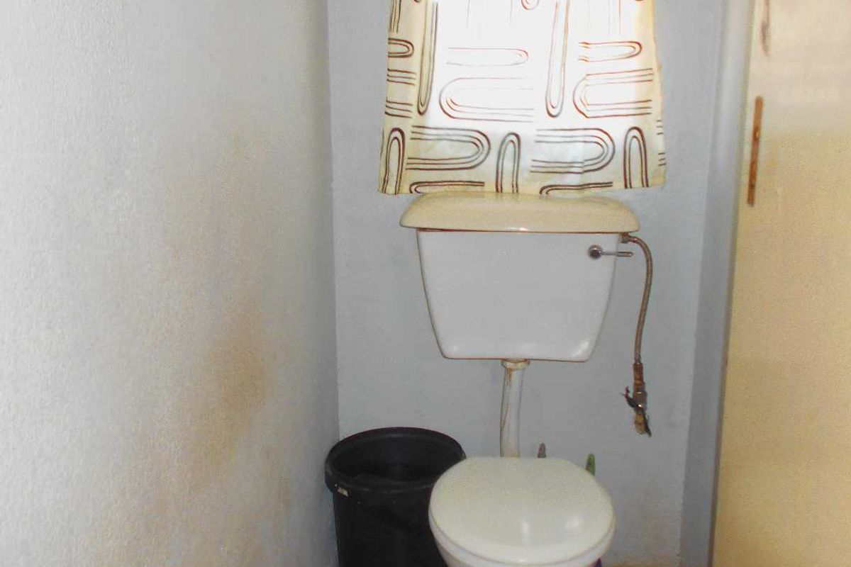 A small, dated bathroom featuring a standard toilet and tiled flooring, currently showing signs of wear and wall staining.