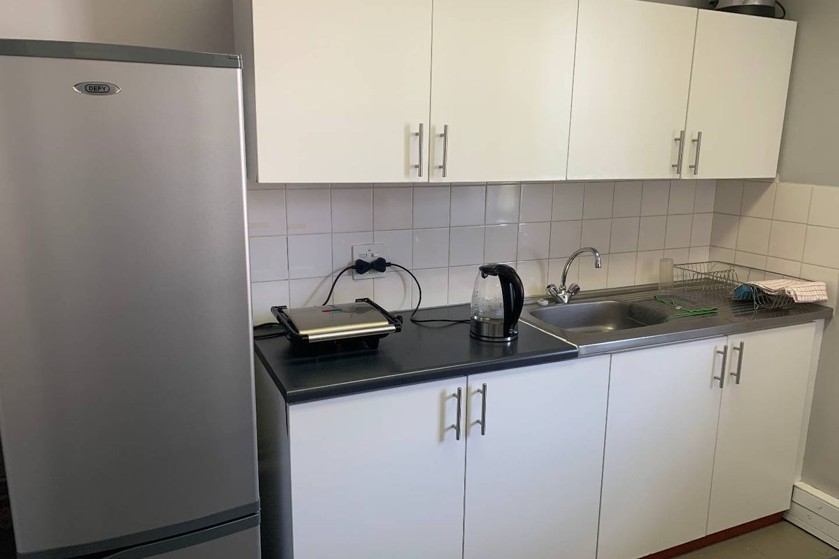 A compact kitchenette featuring white cabinetry, a stainless steel sink, tiled backsplash, and dark laminate countertops.