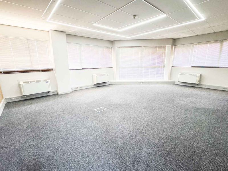 A bright, open-plan office space featuring fitted carpeting, suspended ceiling with recessed lighting, and multiple wall-mounted HVAC units for climate control.