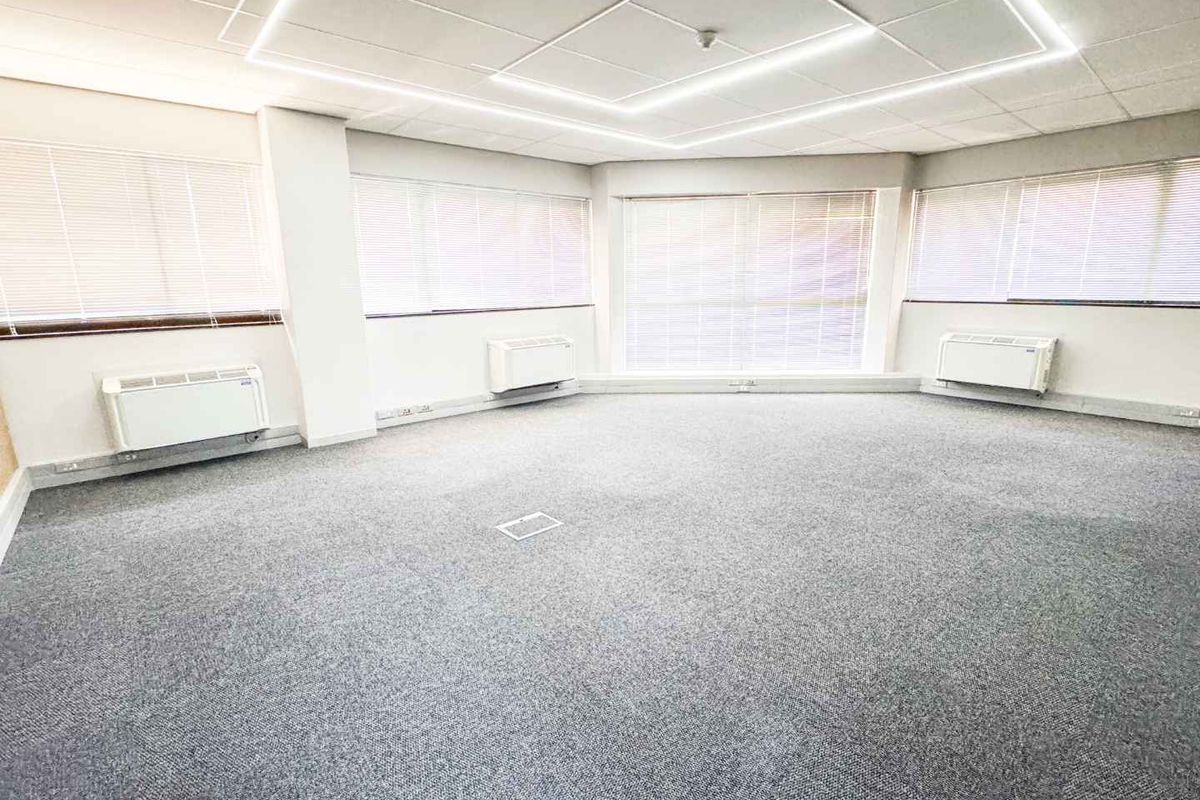 A bright, open-plan office space featuring fitted carpeting, suspended ceiling with recessed lighting, and multiple wall-mounted HVAC units for climate control.