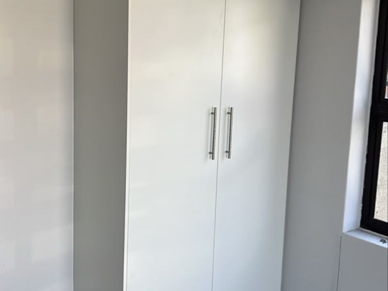 A clean, modern bedroom featuring a white built-in cupboard, neutral tiled flooring, and a window with a dark frame.