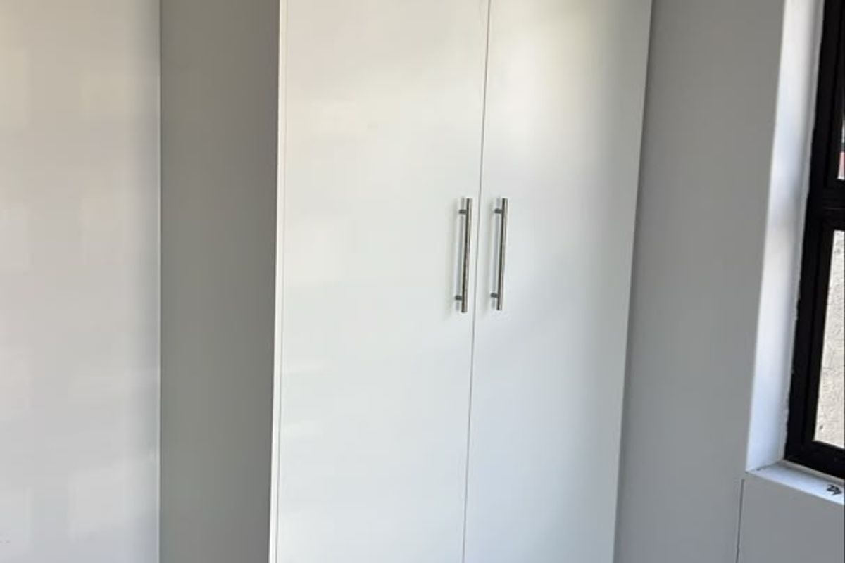 A clean, modern bedroom featuring a white built-in cupboard, neutral tiled flooring, and a window with a dark frame.