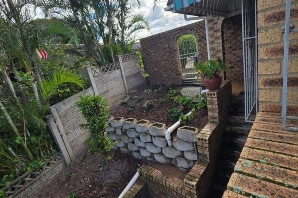 A rear exterior view featuring a multi-level brick garden area with retaining walls, a staircase, and a striped metal awning over the entrance.