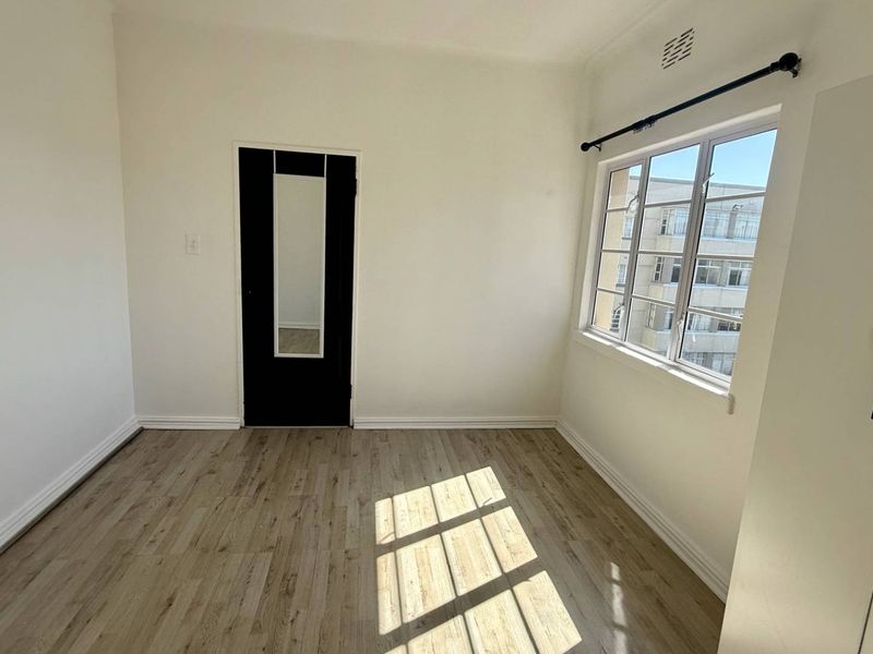 A bright, empty bedroom featuring light-toned laminate flooring, a built-in cupboard, and a window with security bars.