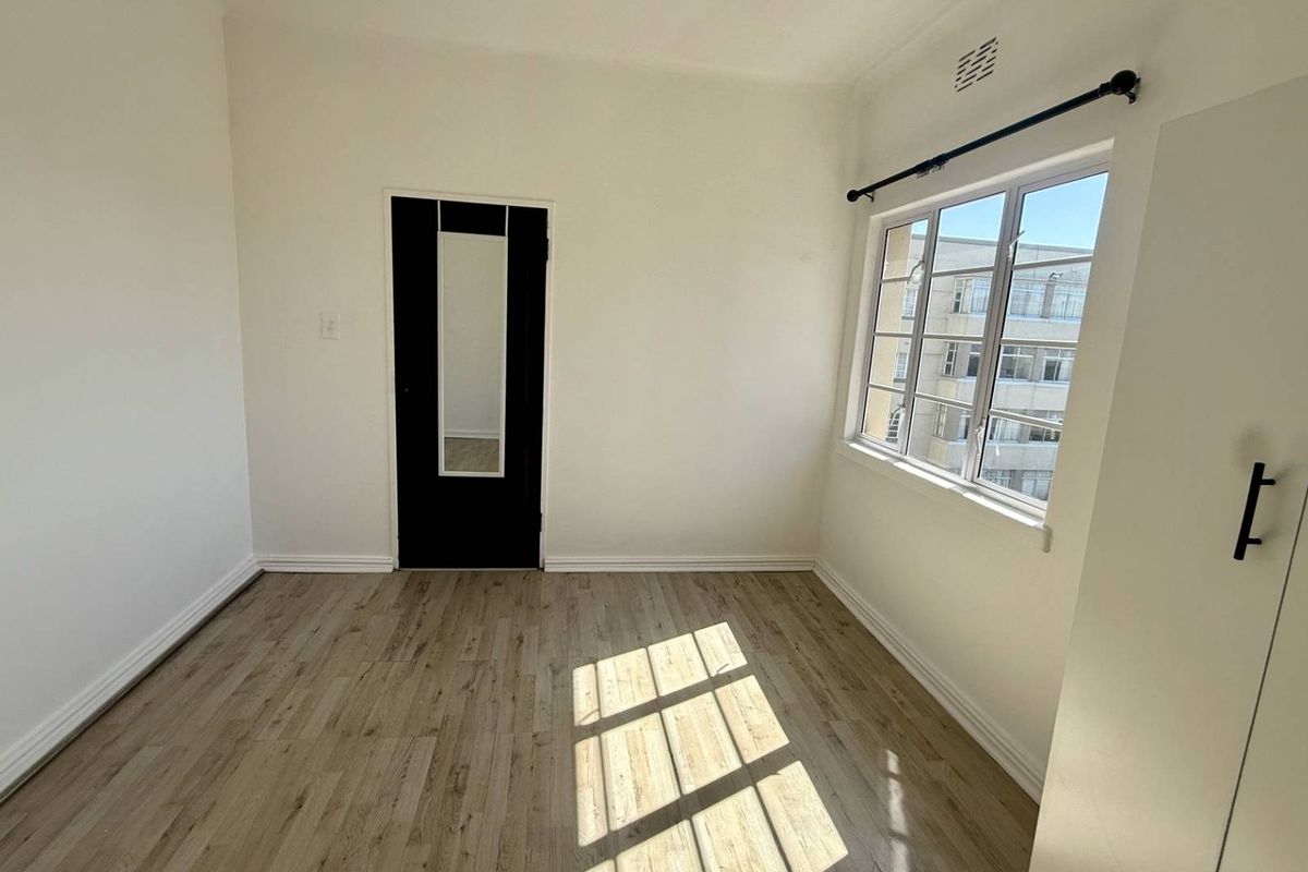 A bright, empty bedroom featuring light-toned laminate flooring, a built-in cupboard, and a window with security bars.