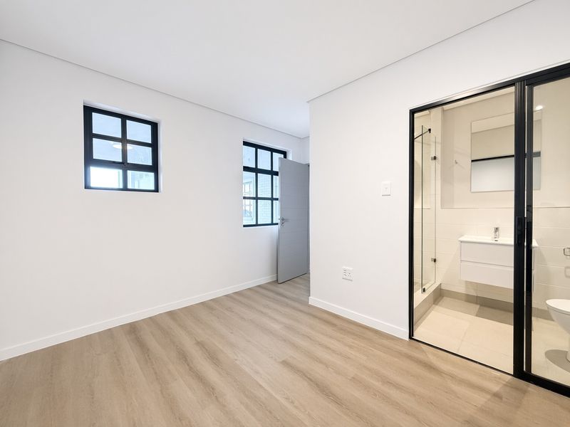 Modern, bright bedroom featuring light laminate flooring, black-framed windows, and a sleek en-suite bathroom with a glass-enclosed shower and wall-mounted vanity.