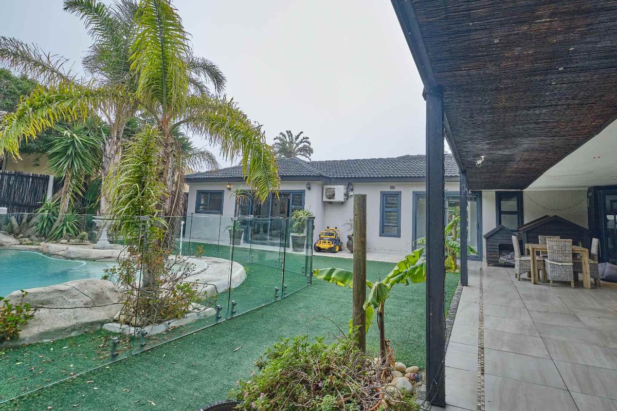 Rear exterior view featuring a covered tiled patio, artificial lawn, and a swimming pool with glass safety fencing.
