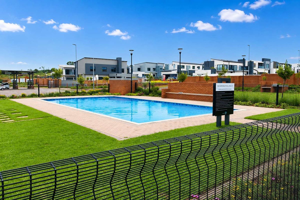A well-maintained communal swimming pool area featuring paved surrounds, brick boundary walls, and secure mesh fencing within a residential estate.