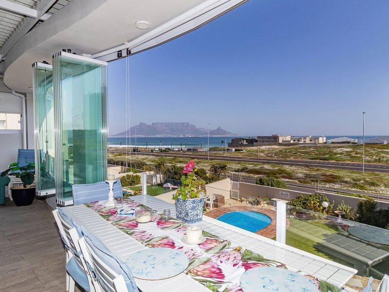 Spacious covered patio featuring stacking glass doors, tiled flooring, and a glass balustrade, offering panoramic views of the coastline and Table Mountain.