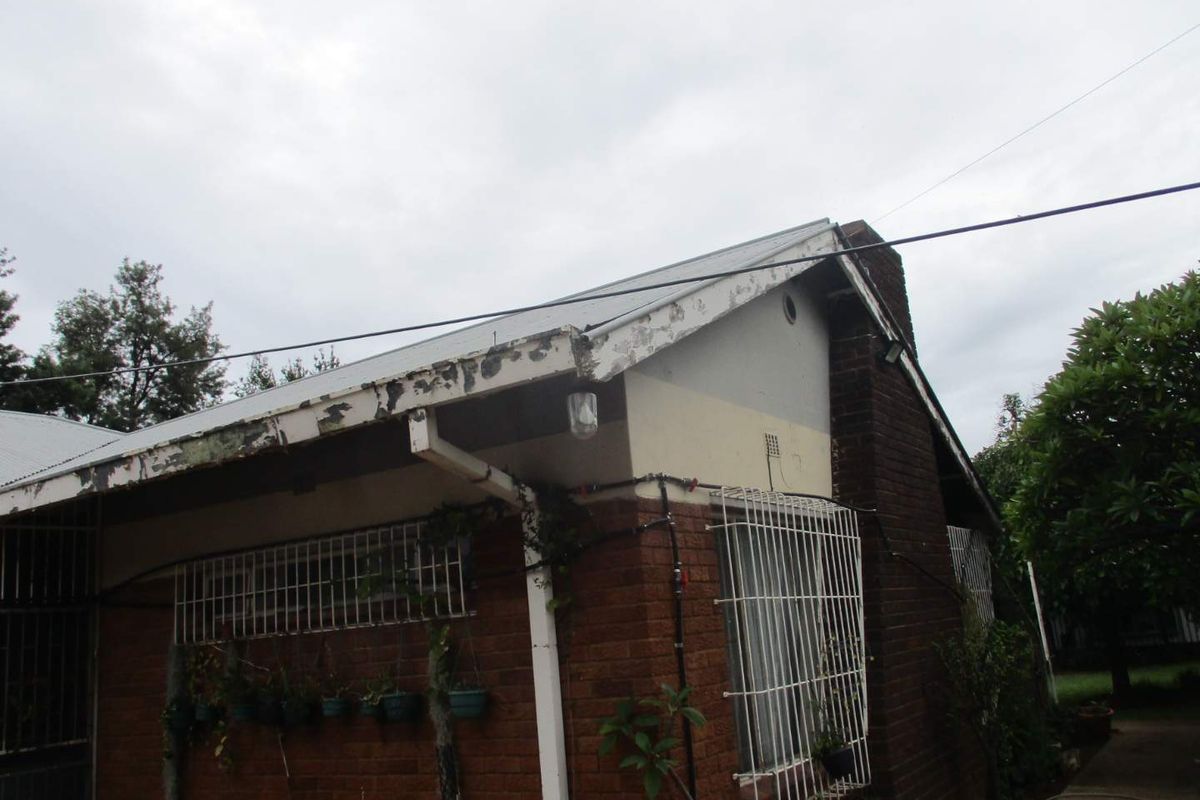 Rear exterior view of the house featuring brick construction, a metal roof, and security bars on windows, showing signs of exterior maintenance required.