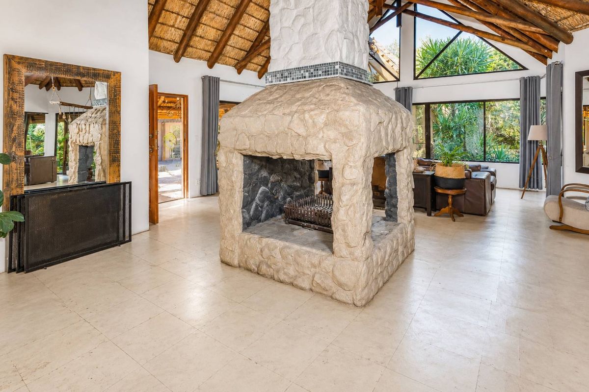 Spacious living area featuring a prominent central stone fireplace, exposed timber trusses, high thatched ceilings, and large windows providing ample natural light.