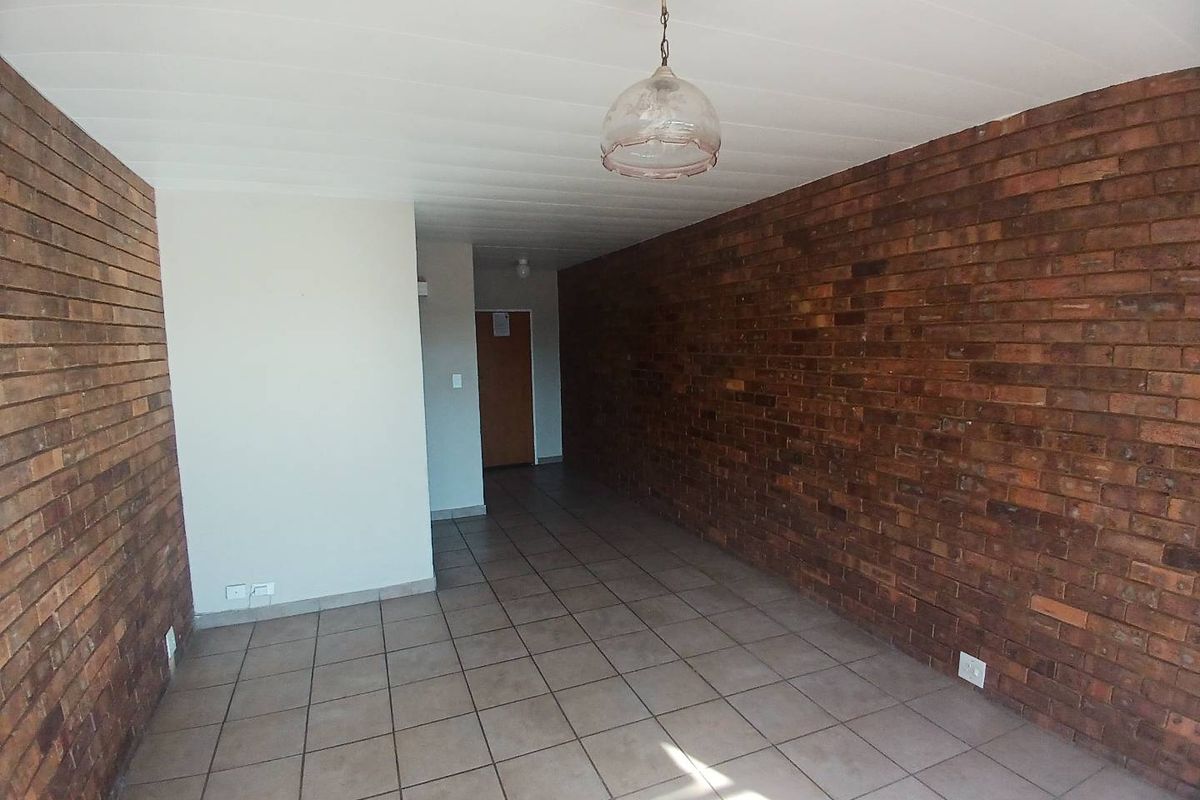 An empty living area featuring exposed brick walls, tiled flooring, and a central pendant light fixture, offering a blank canvas for renovation.