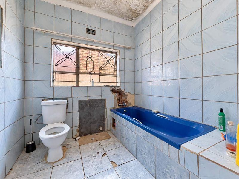 A bathroom in need of significant renovation, featuring a blue bathtub, toilet, and damaged wall and floor tiling throughout the space.