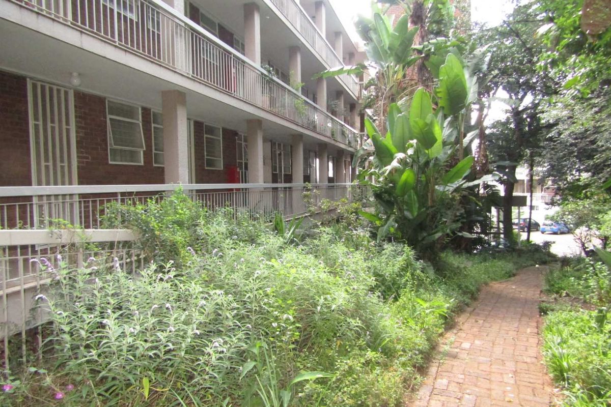 Exterior view of the apartment building featuring a brick facade, multi-level balconies with metal railings, and a paved garden walkway.