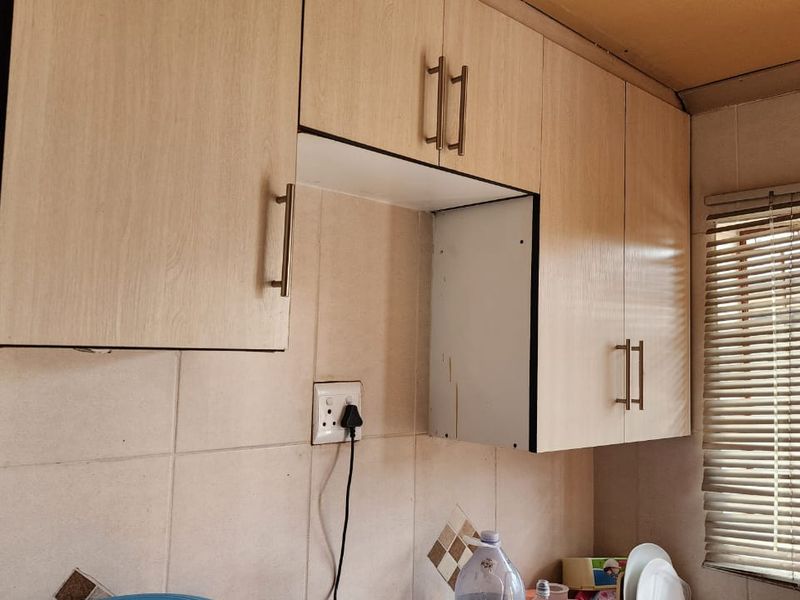 A kitchen area featuring light wood-look laminate cabinetry, tiled backsplash, and venetian blinds, showing signs of wear on the cabinet finishes.