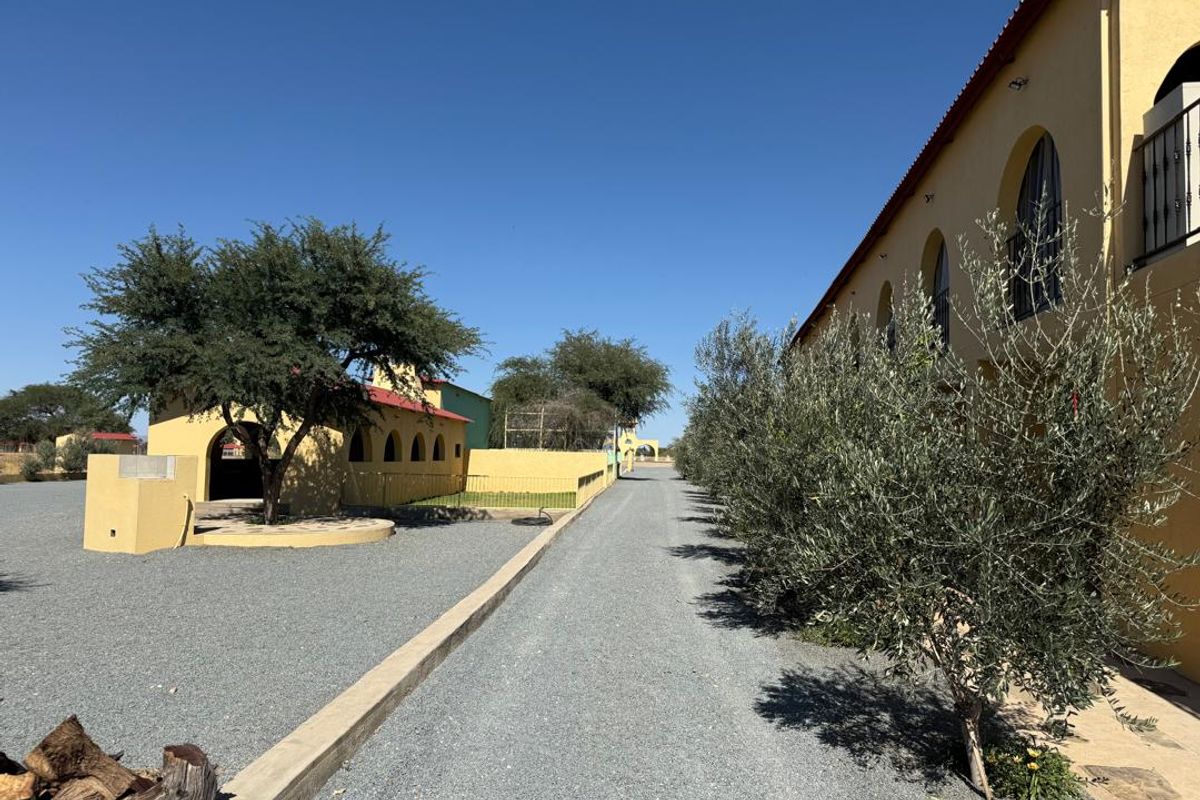 A view of the property grounds featuring a gravel driveway, landscaped trees, and yellow building facades with arched architectural details.