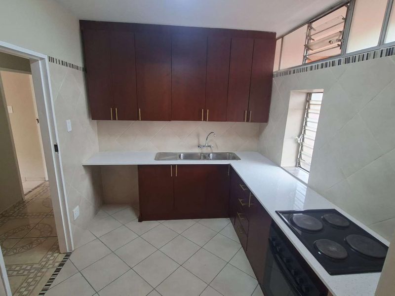 A clean, functional kitchen featuring dark wood cabinetry, white countertops, a built-in electric hob and oven, and tiled finishes throughout.