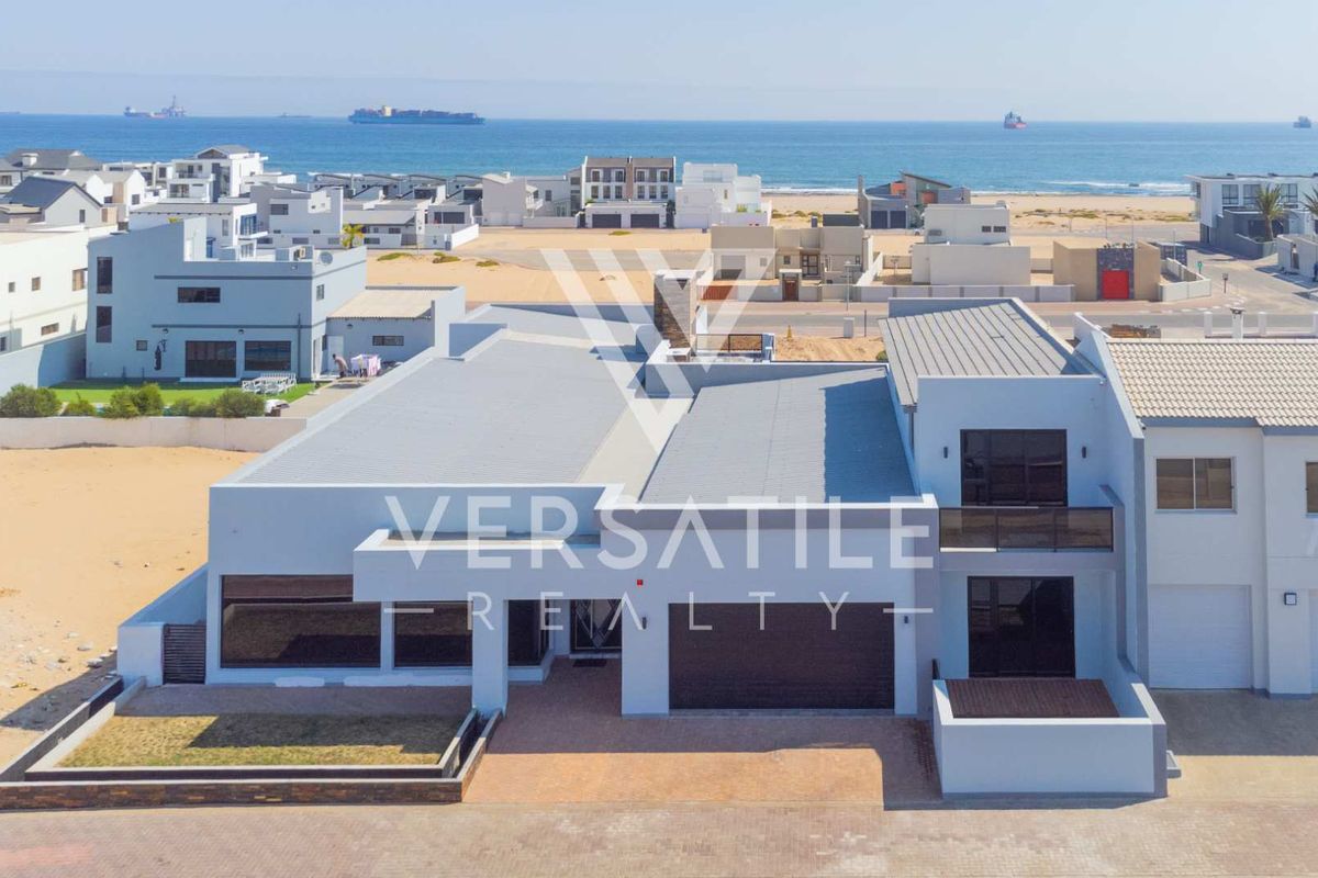 Aerial view of a modern coastal residence featuring a metal roof, paved driveway, and balcony, situated in a prime seaside location.