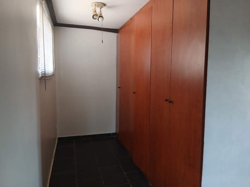 A functional bedroom space featuring dark tiled flooring, built-in wooden cupboards, and a window with venetian blinds.