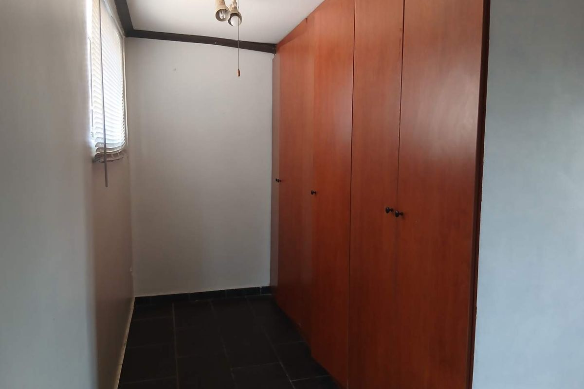 A functional bedroom space featuring dark tiled flooring, built-in wooden cupboards, and a window with venetian blinds.