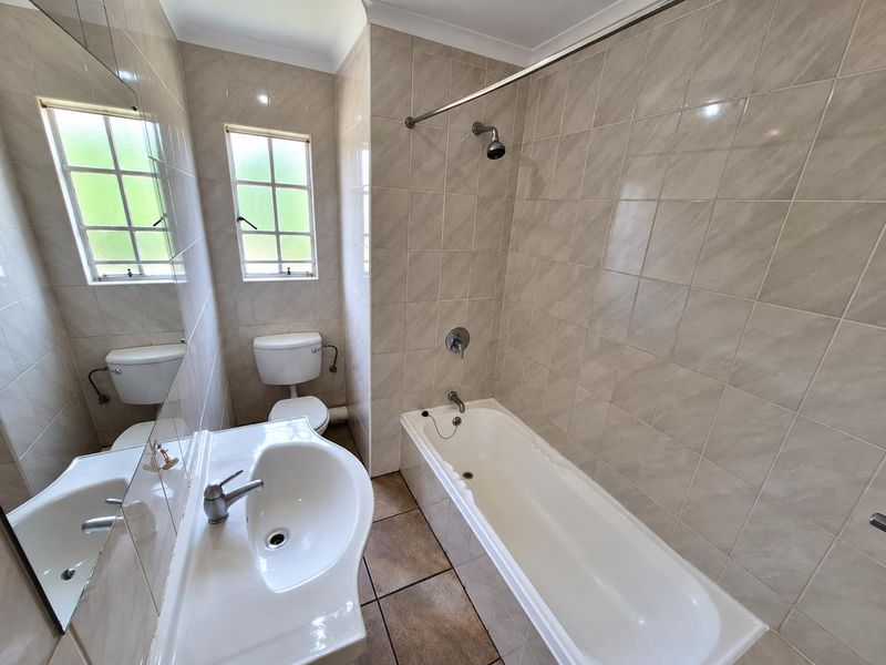 A functional bathroom featuring a built-in bathtub with shower, pedestal basin, and toilet, finished with neutral wall and floor tiling.