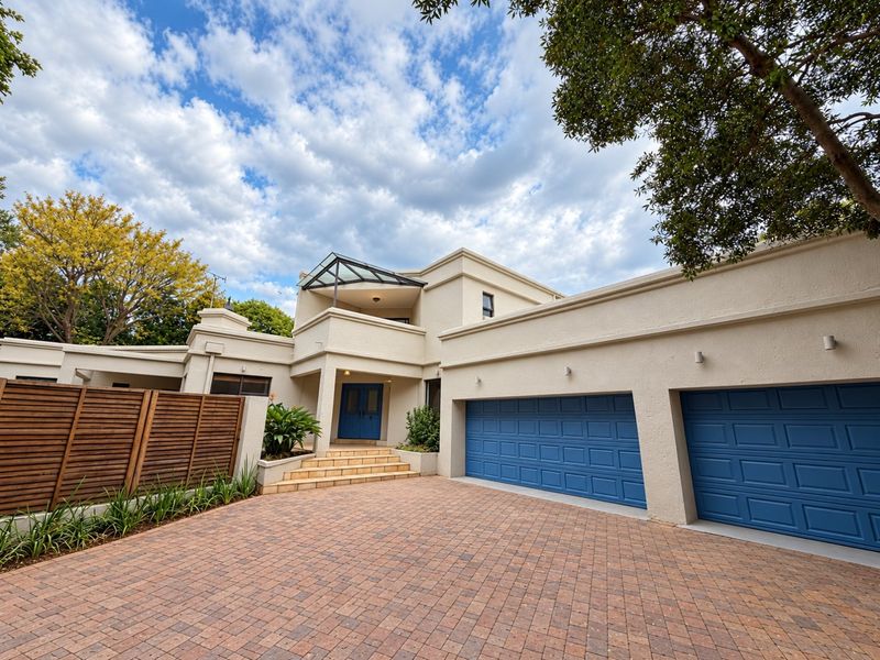 A well-maintained front exterior featuring a paved driveway, double garage with blue doors, and a covered entrance with a glass canopy.