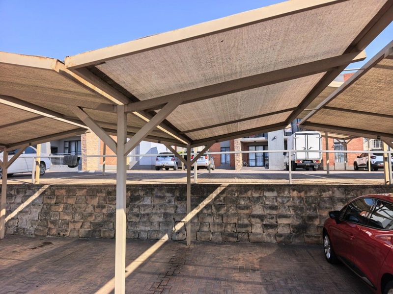 Covered carport area featuring a steel frame with shade cloth covering, situated on a paved surface with a stone retaining wall.