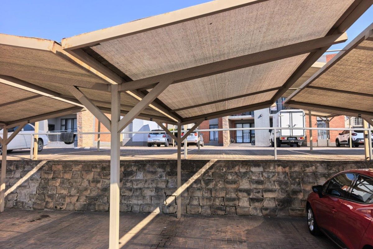 Covered carport area featuring a steel frame with shade cloth covering, situated on a paved surface with a stone retaining wall.