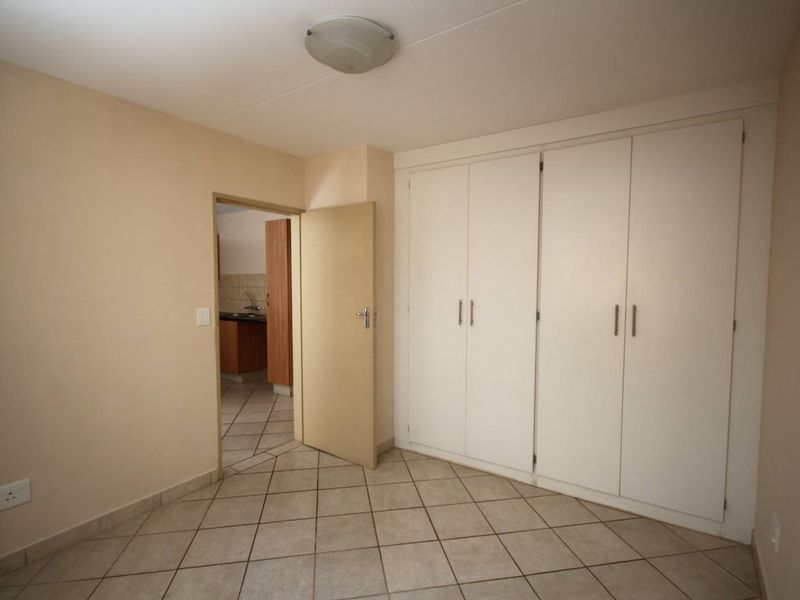 A clean, empty bedroom featuring neutral tiled flooring and ample built-in cupboard storage, with a doorway leading to an adjacent kitchen area.