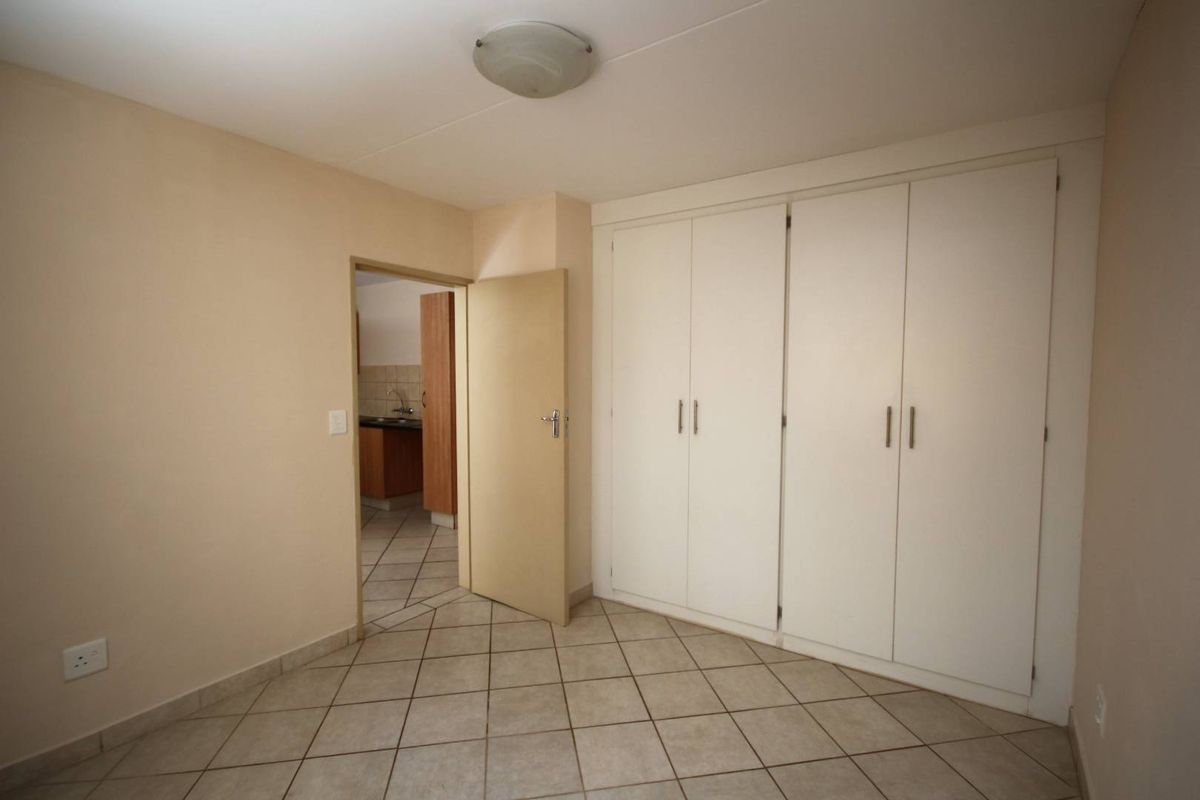 A clean, empty bedroom featuring neutral tiled flooring and ample built-in cupboard storage, with a doorway leading to an adjacent kitchen area.