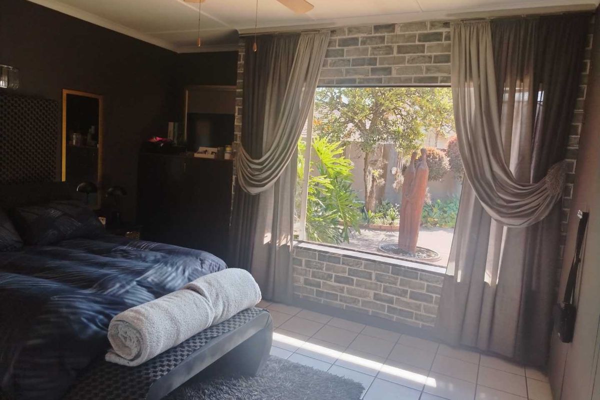 A well-lit bedroom featuring an exposed brick accent wall, large window with garden views, ceiling fan, and neutral tiled flooring.
