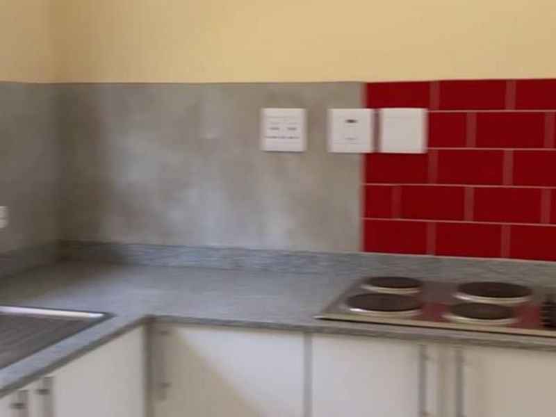 A compact kitchen featuring white cabinetry, stone countertops, a stainless steel sink, and an electric hob with a partially tiled splashback.