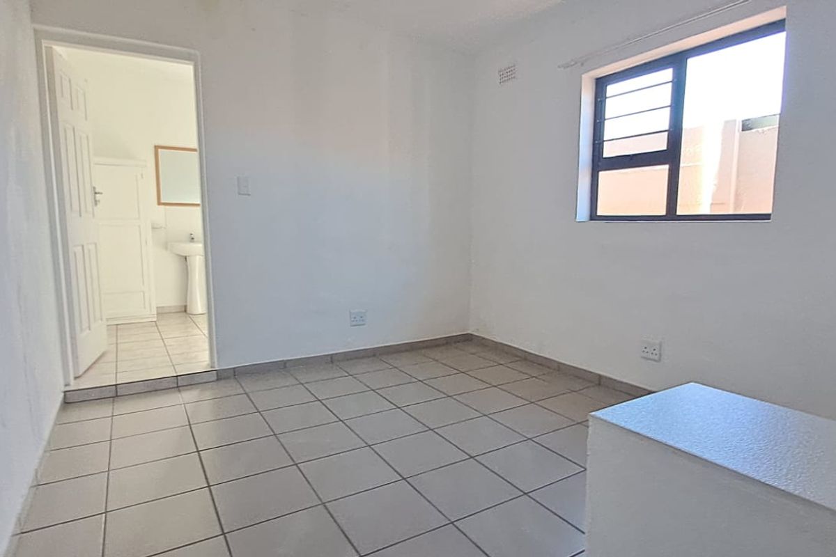 A bright, empty bedroom featuring neutral floor tiles, a window with security bars, and direct access to an adjoining bathroom.