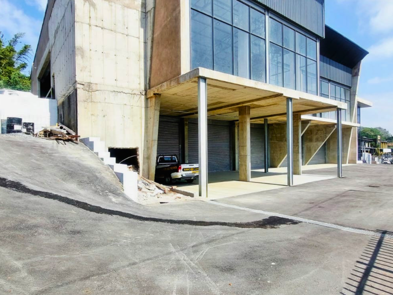 Industrial property exterior featuring a concrete facade, large glass windows, multiple roller shutter doors, and an asphalt driveway with ramp access.