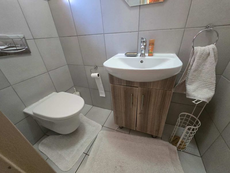 A compact, modern bathroom featuring a wall-mounted toilet, a wood-finish vanity cabinet with a ceramic basin, and neutral wall and floor tiling.