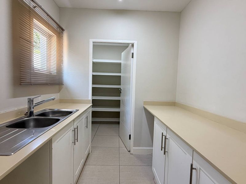 A clean and functional scullery featuring double bowl sink, ample built-in cabinetry, solid surface countertops, and a convenient walk-in pantry.