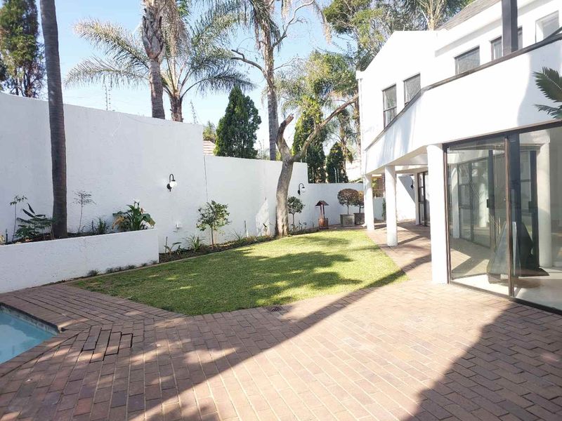 Rear exterior view featuring a paved patio area, a swimming pool, a manicured lawn, and raised garden beds against a high boundary wall.
