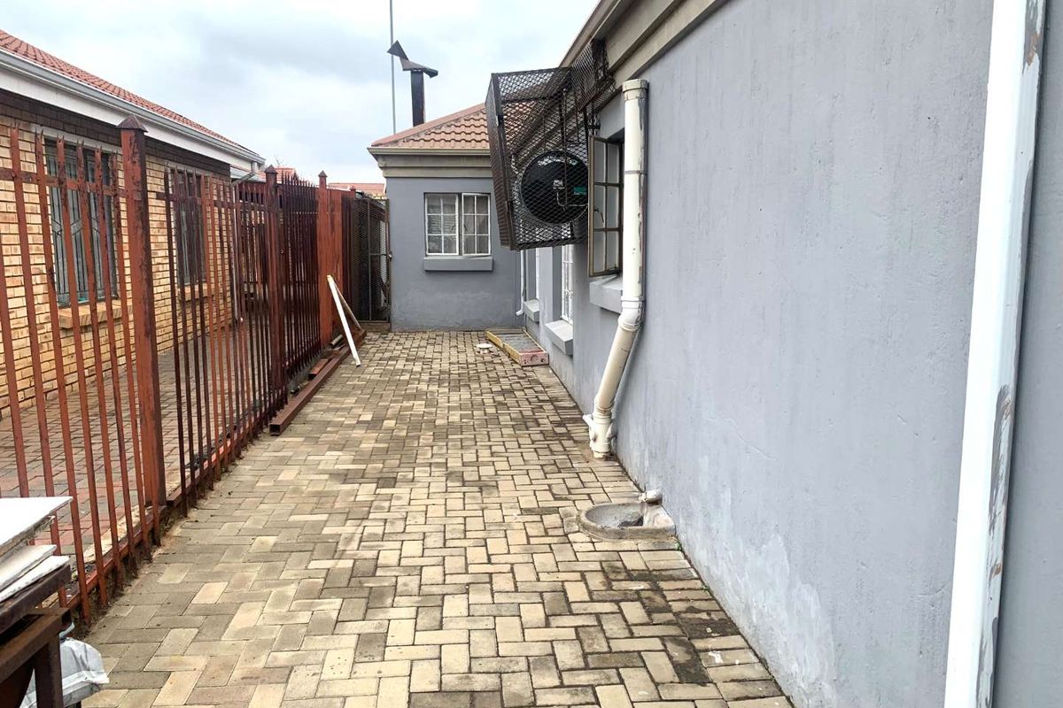 A narrow paved side passage featuring a metal security fence, external air conditioning unit, and drainage infrastructure along the exterior wall.