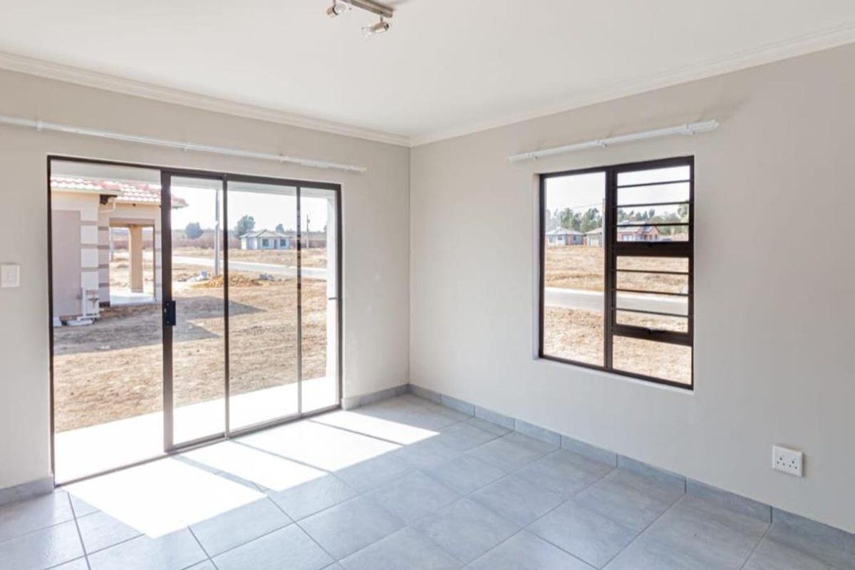 A bright, empty living room featuring neutral tiled flooring, large sliding glass doors leading to the exterior, and a window with dark frames.