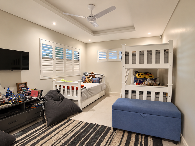 Spacious bedroom featuring a tray ceiling with a fan, recessed lighting, and plantation shutters, finished with neutral tiled flooring.