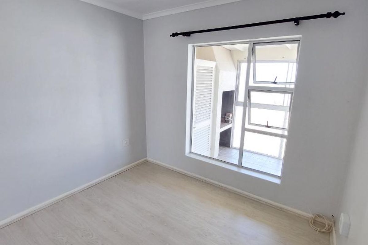A bright, empty bedroom featuring light-toned laminate flooring, a ceiling light fixture, and a window overlooking an outdoor patio area.