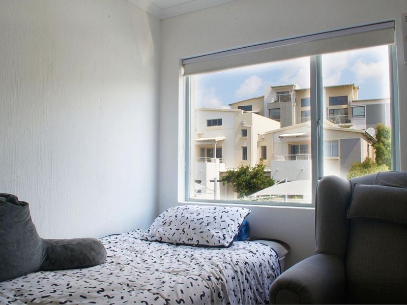 A bedroom featuring a large window with a roller blind, offering natural light and a view of the surrounding apartment complex.