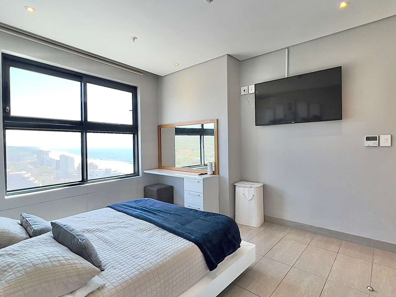 A bright, modern bedroom featuring large windows with coastal views, tiled flooring, recessed lighting, and a built-in vanity area.