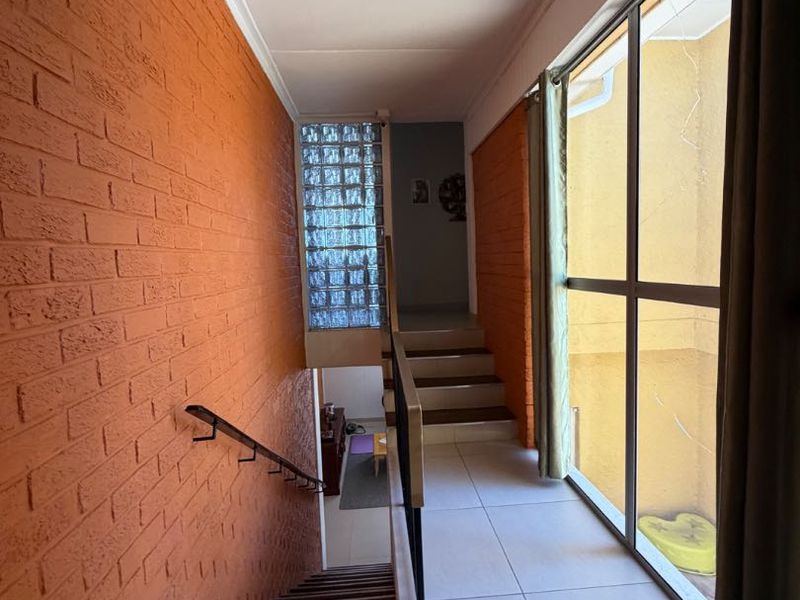 A bright hallway featuring a distinctive orange brick wall, tiled flooring, and a staircase with a metal handrail, illuminated by a large glass window.