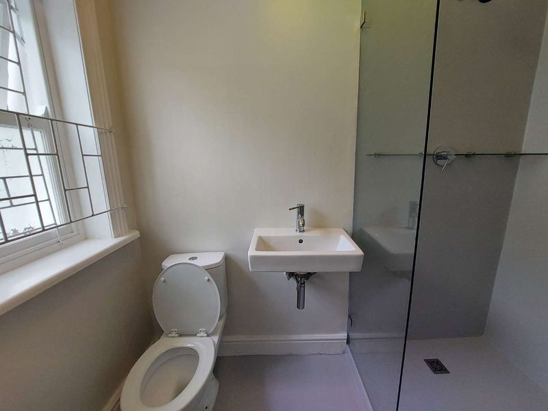A functional bathroom featuring a wall-mounted basin, a toilet, and a glass-enclosed shower, illuminated by a classic leaded window.